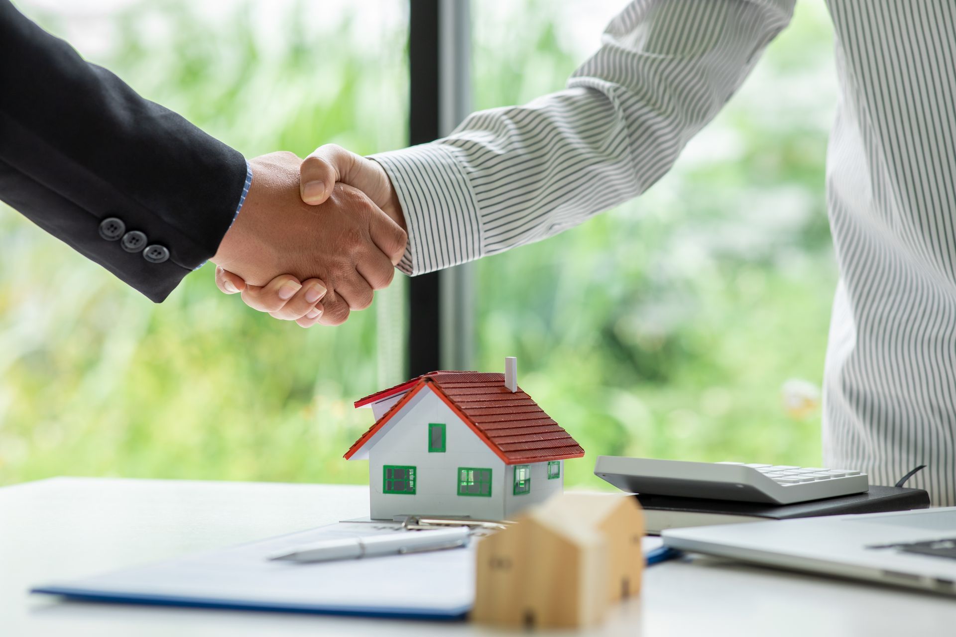 Two people shaking hands over a miniature house, symbolizing a real estate deal.