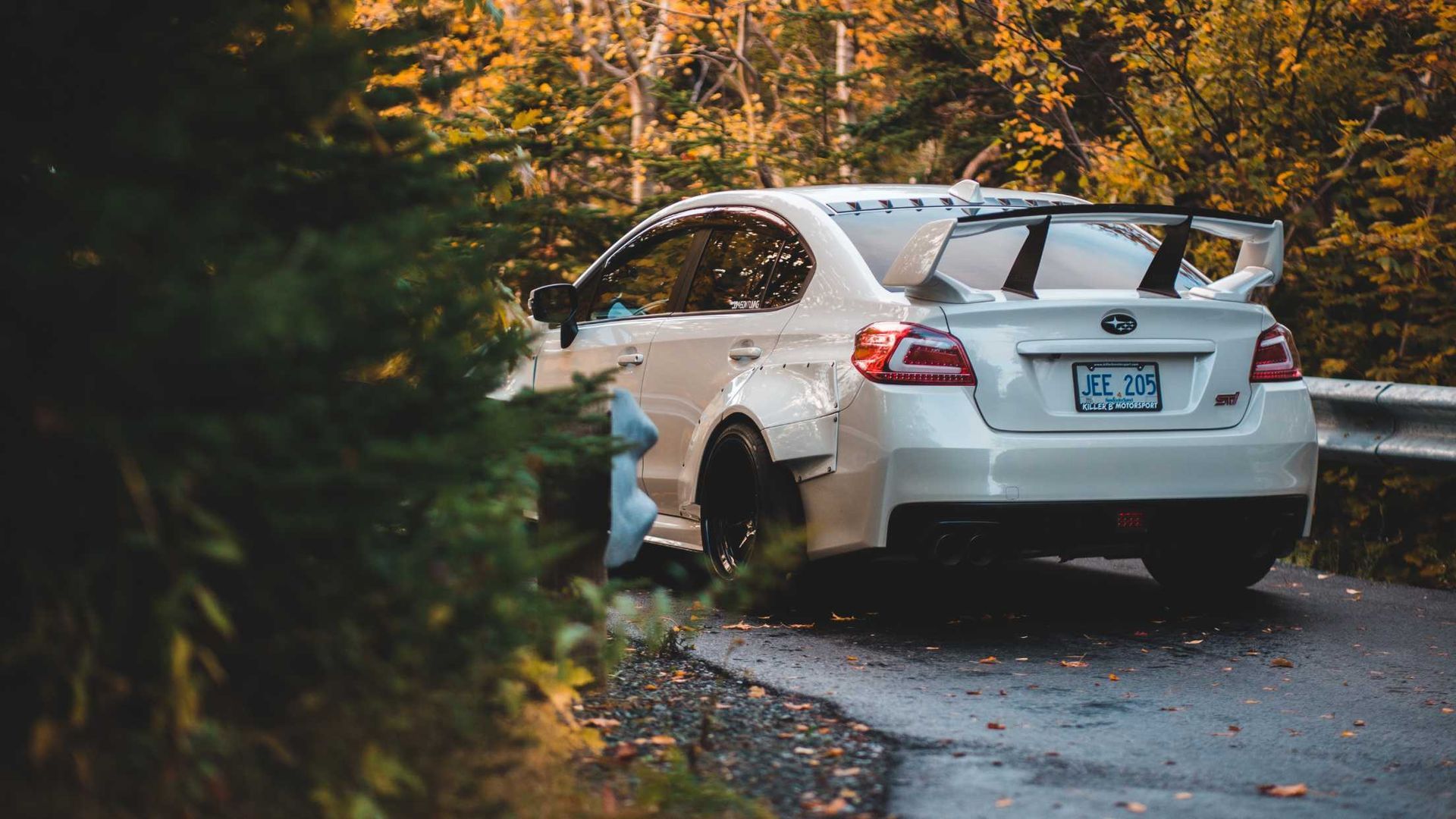 White Subaru sedan with large spoiler on a road, surrounded by autumn foliage.