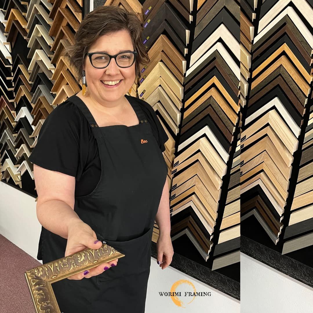 A Woman Is Holding A Picture Frame In Front Of A Wall Of Picture Frames — Worimi Framing Service In Adamstown, NSW