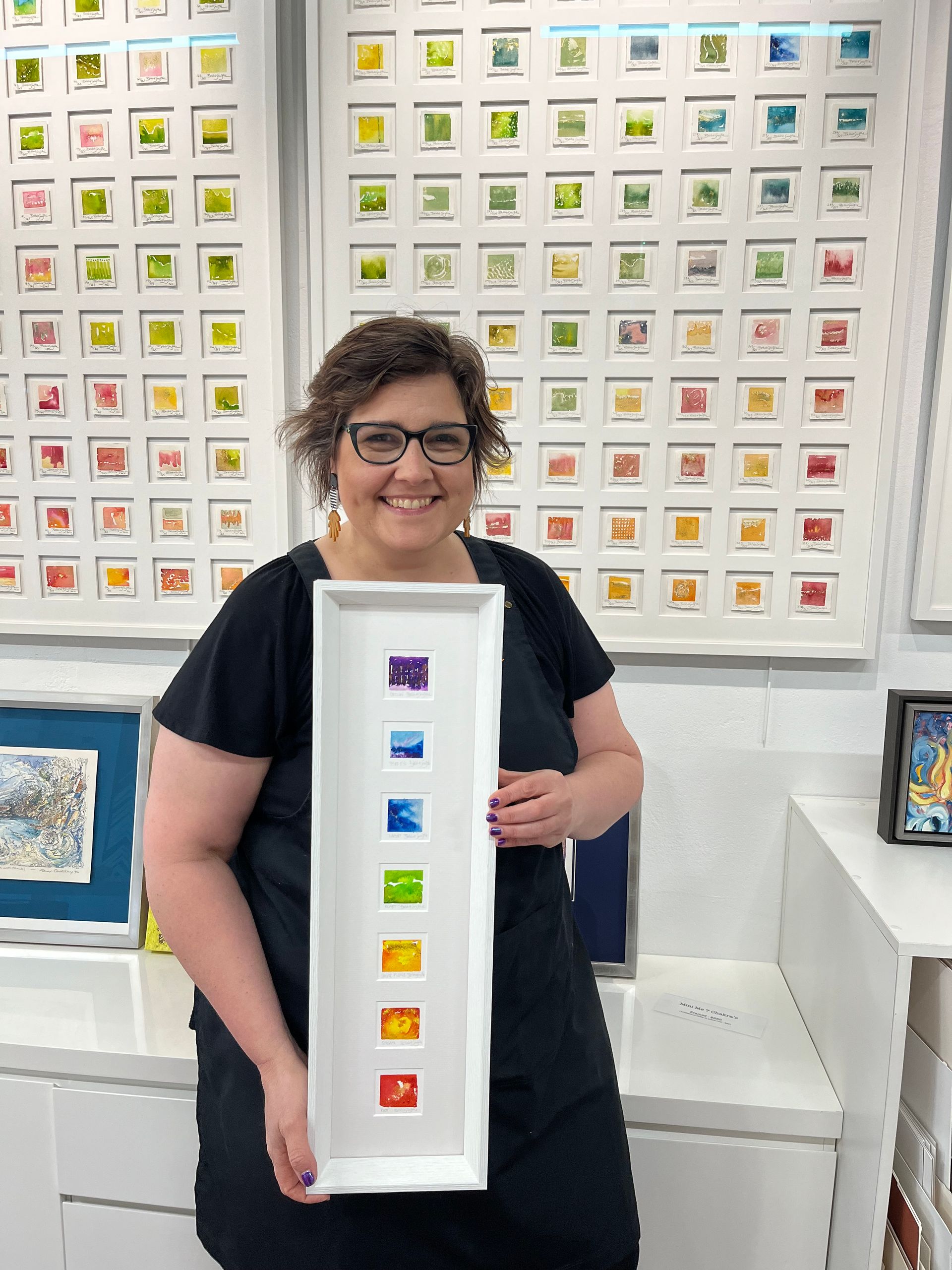 A Woman Holding A Painting Infront Of A Busy Wall Of Paintings— Worimi Framing Service In Adamstown, NSW