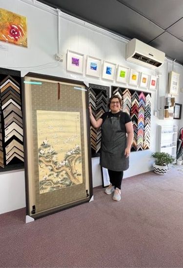 A Woman Is Holding A Large Framed Painting In Front Of A Wall Of Paintings — Worimi Framing Service In Central Coast, NSW
