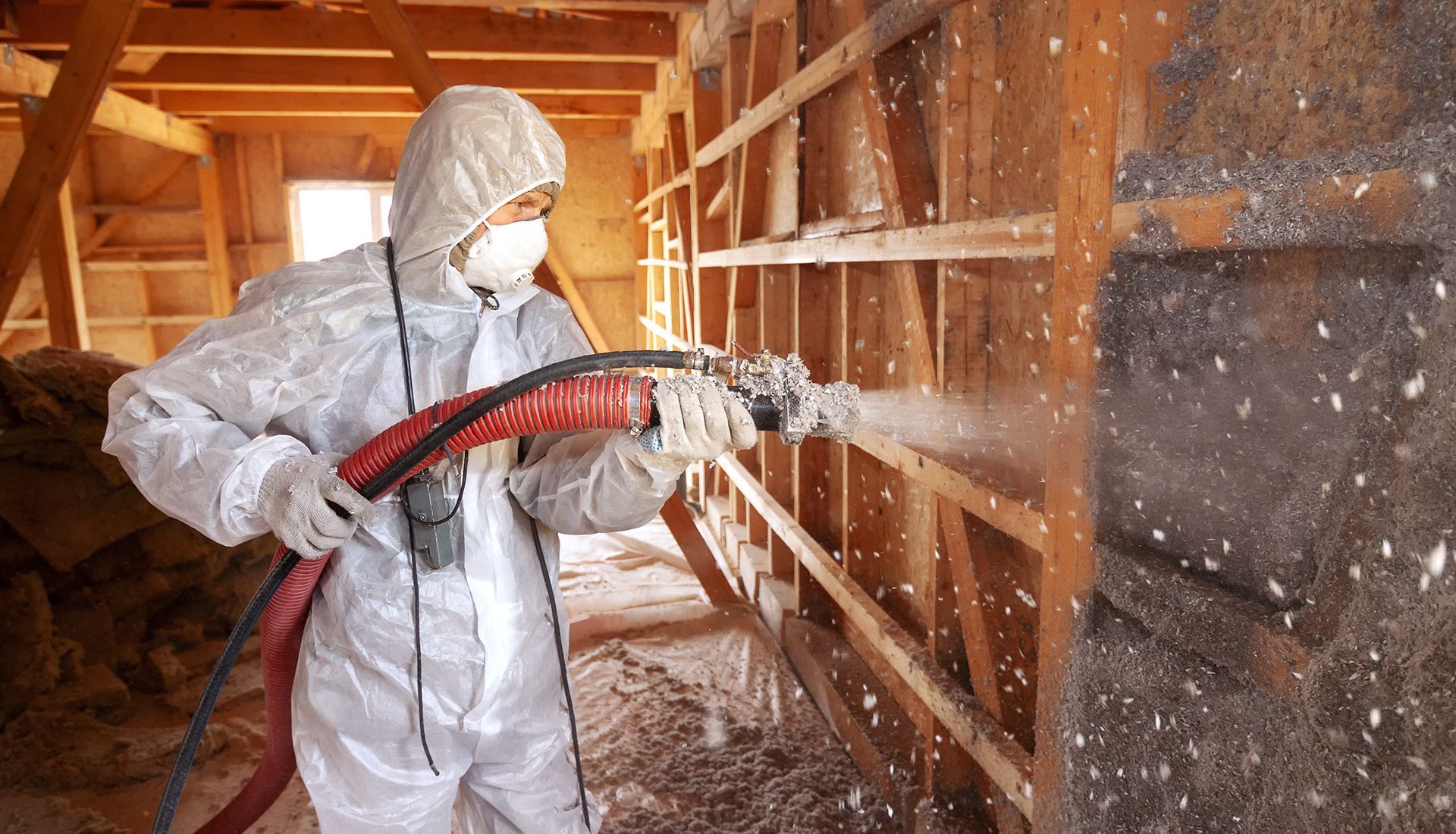 A man in a protective suit is spraying insulation on a wooden wall.