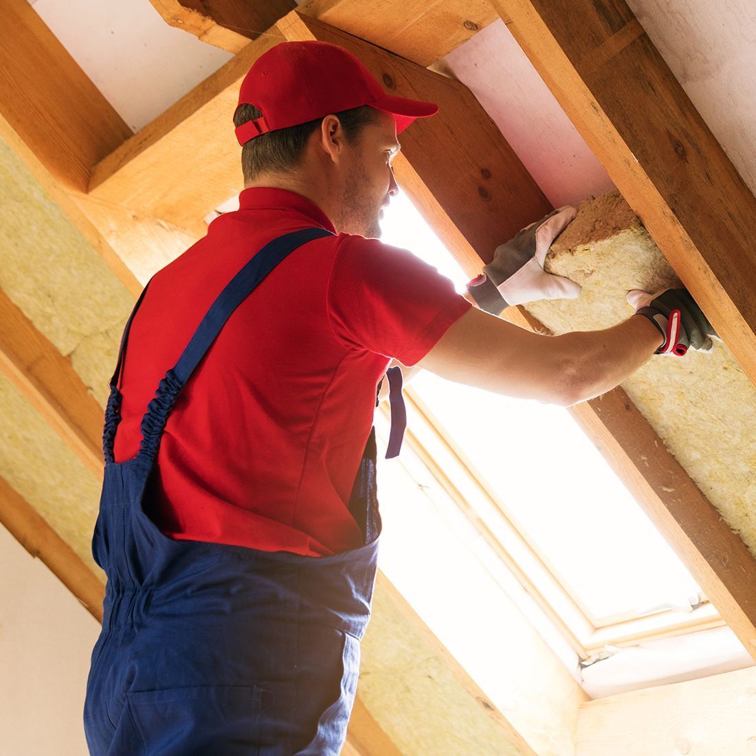 A man in a red shirt and blue overalls is working on a roof.