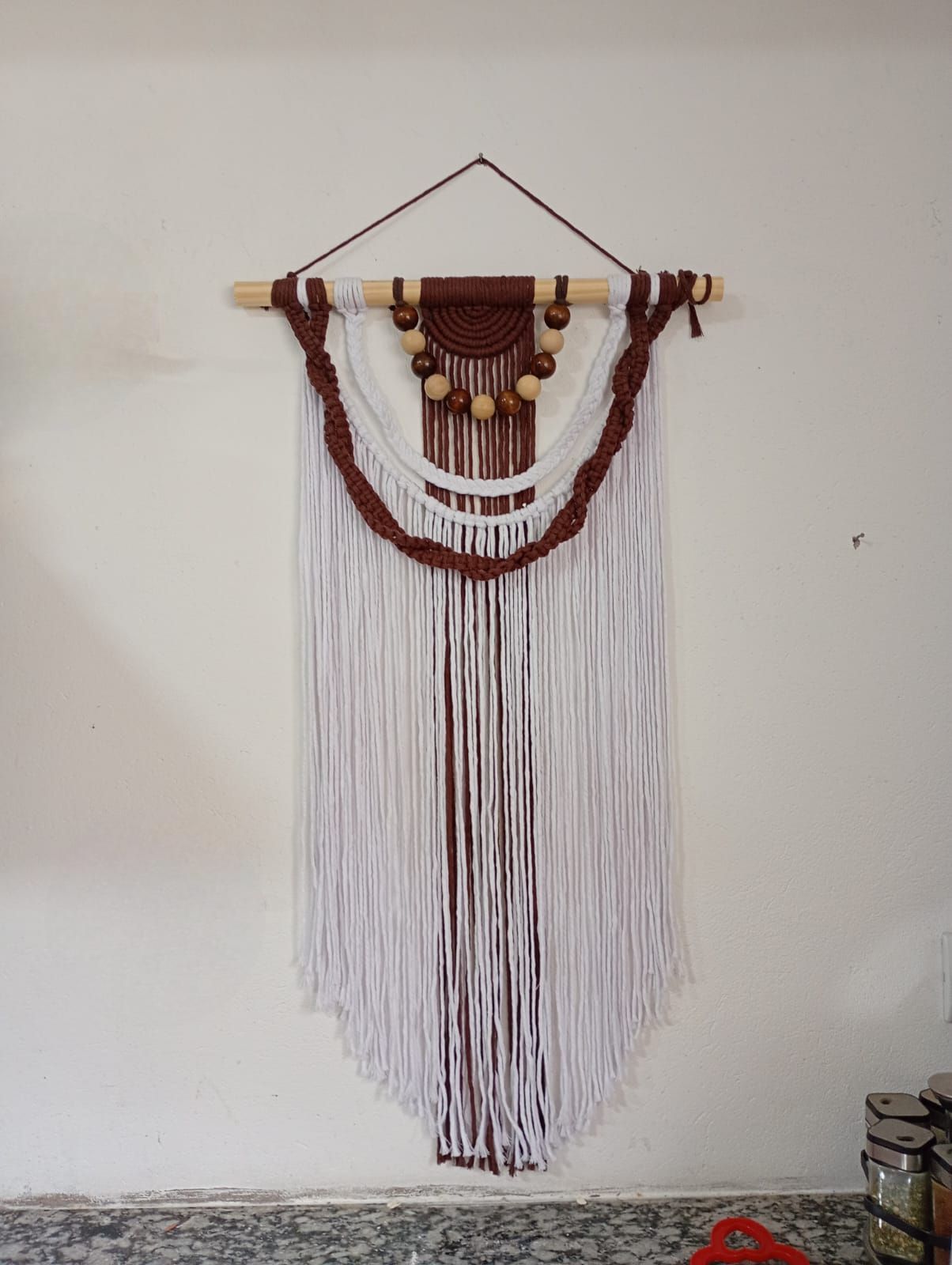 Tapiz de macramé con hilo marrón y blanco, cuentas de madera y una varilla de madera natural.