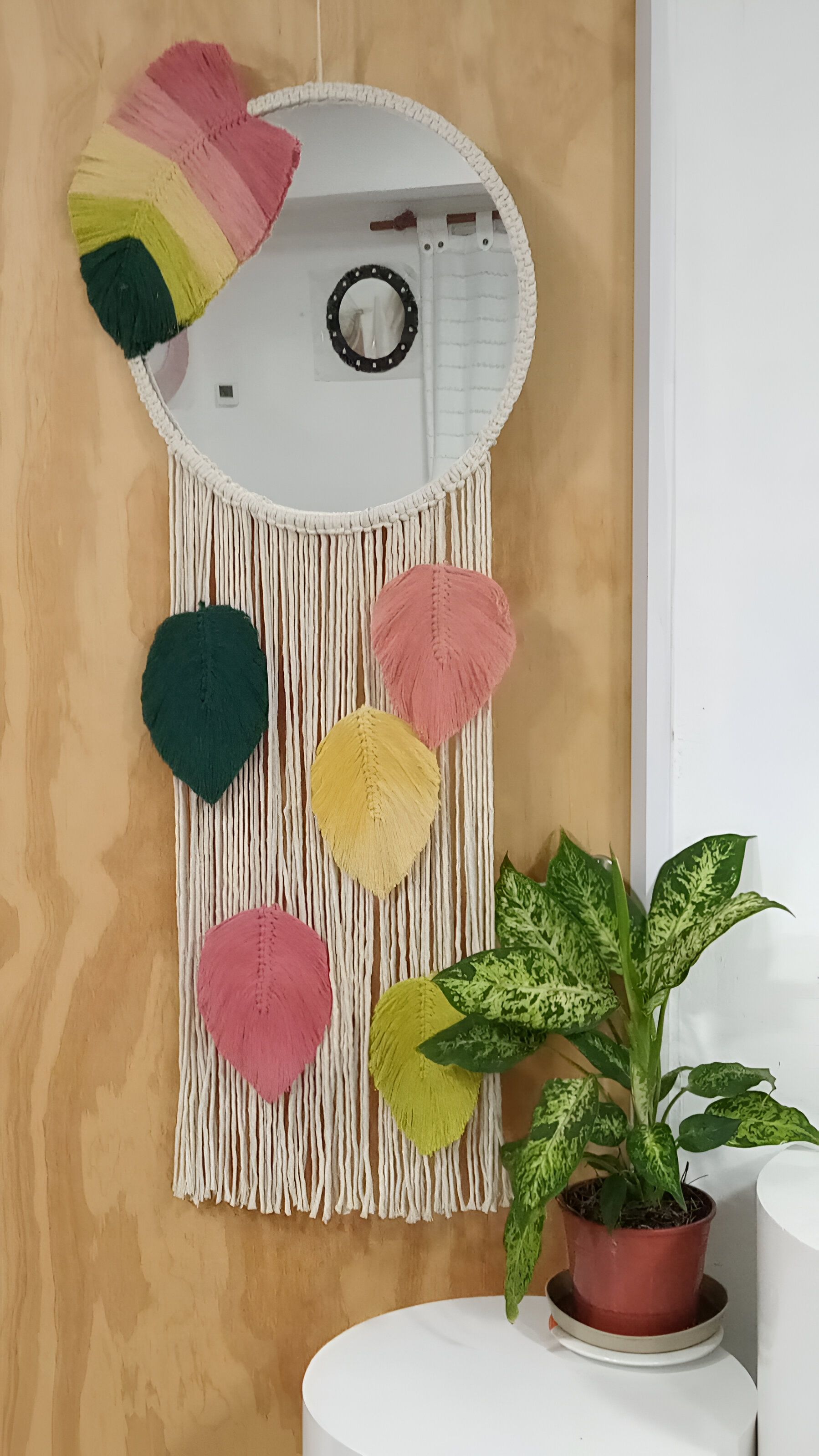 Tapiz de macramé con espejo redondo y decoración de hojas coloridas; planta sobre una mesa auxiliar blanca.