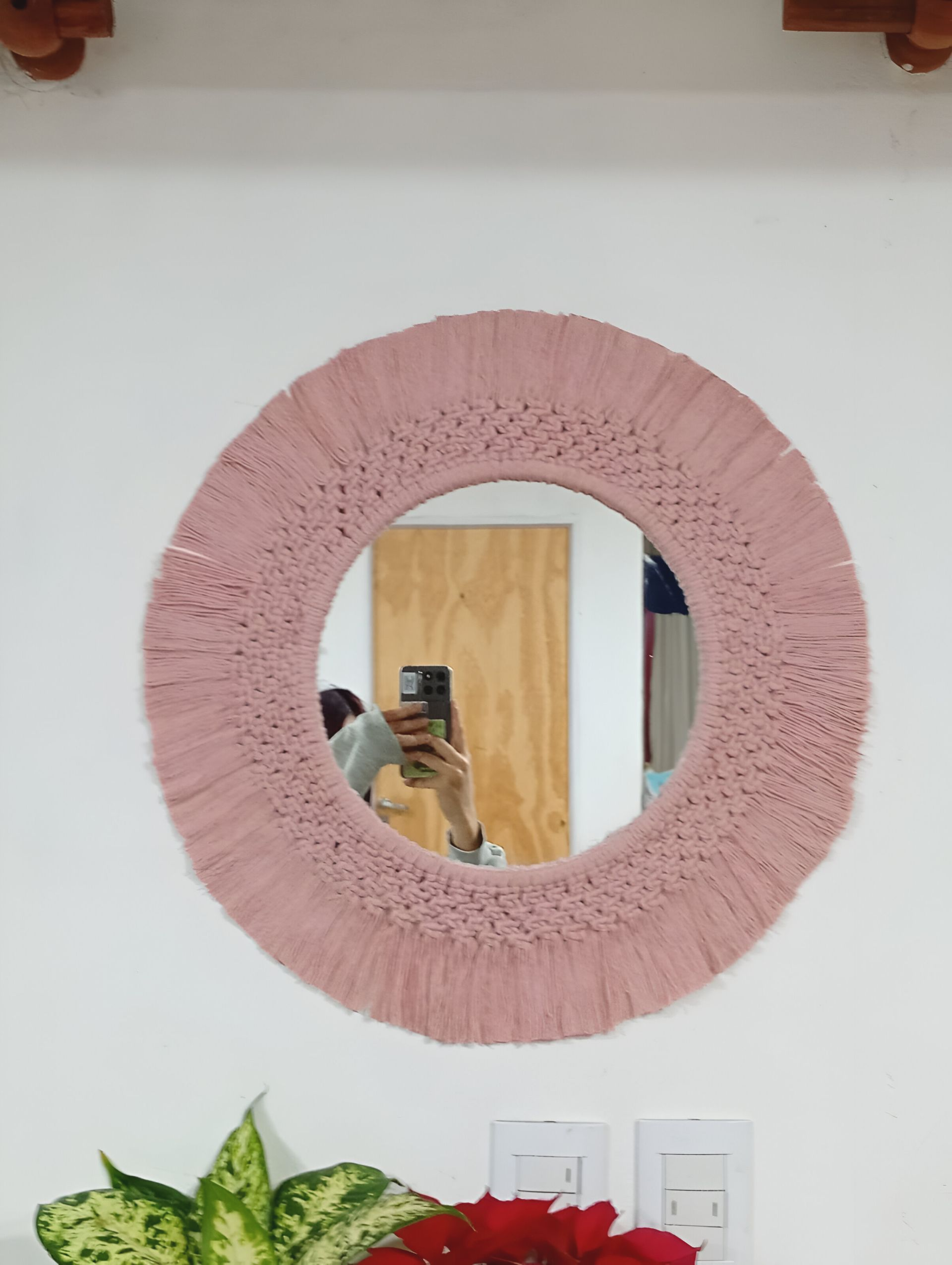 Espejo redondo de macramé rosa sobre pared blanca, con el reflejo de una persona tomando una foto.