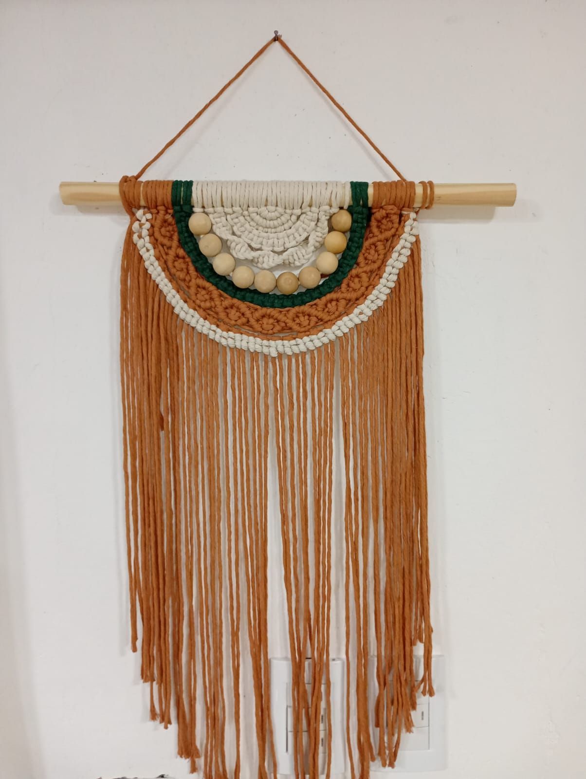 Tapiz de macramé con motivos en tonos beige, blanco y verde, cuentas de madera y flecos largos.