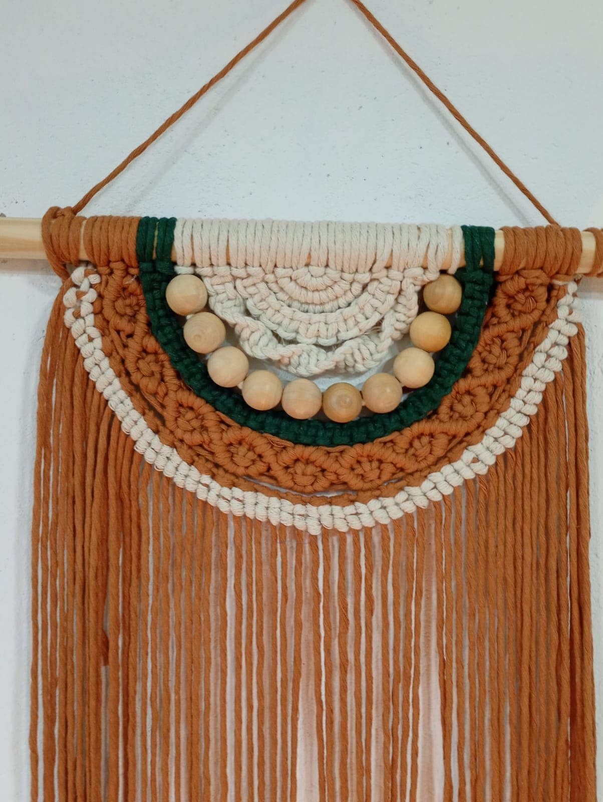 Tapiz de macramé con motivos en tonos beige, blanco y verde, cuentas de madera y flecos largos.