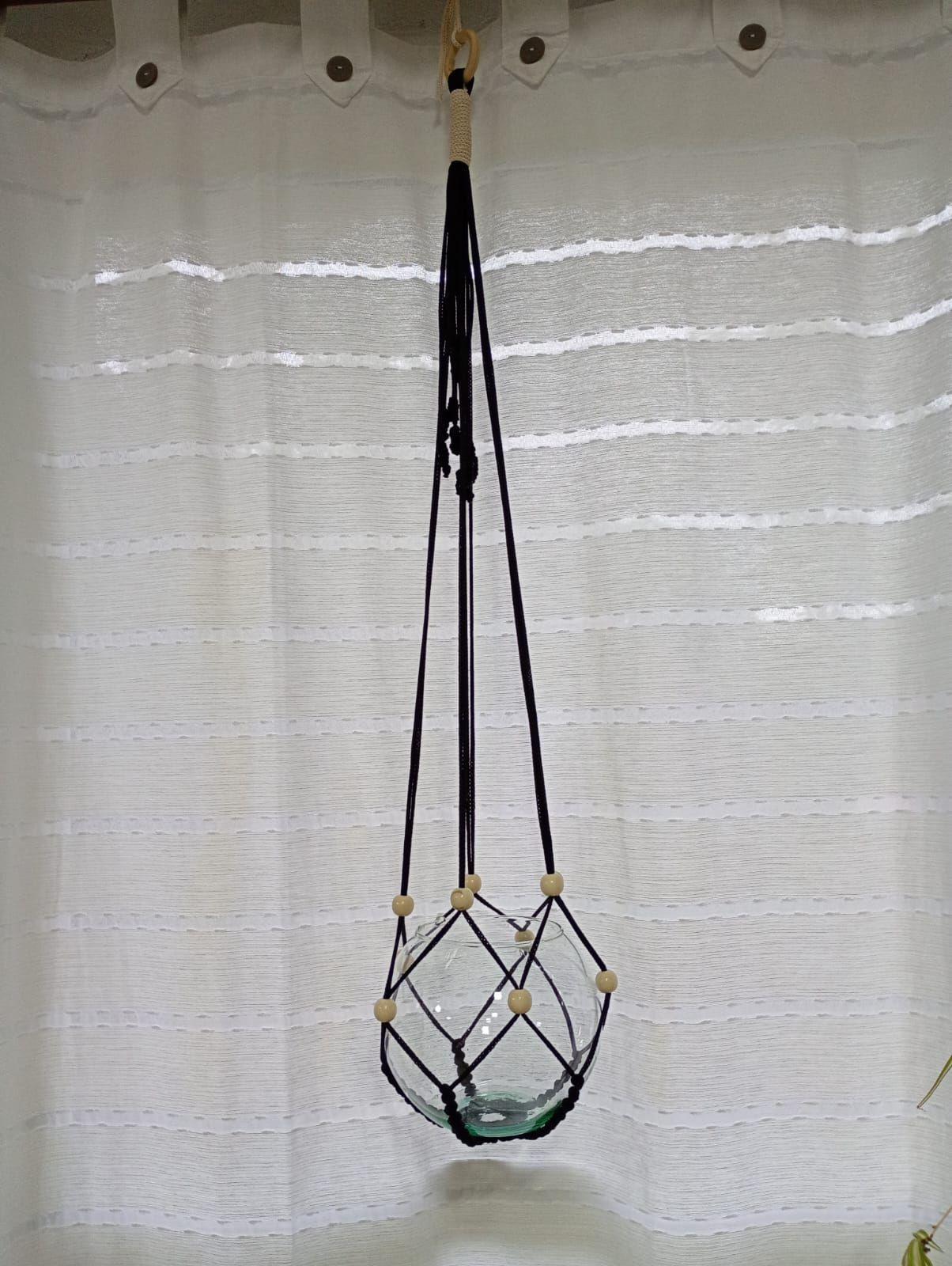 Macetero colgante de macramé negro que sostiene un cuenco de cristal transparente, colgado contra una cortina blanca.