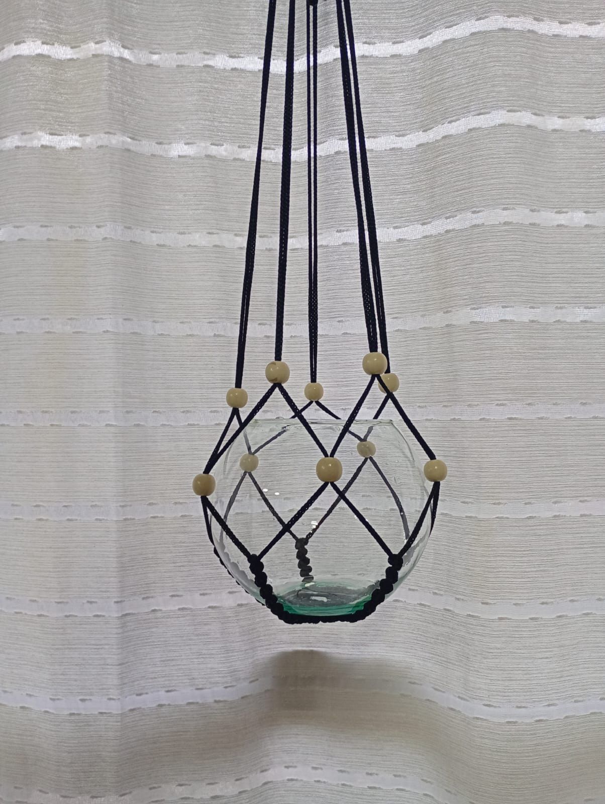 Macetero colgante de macramé negro que sostiene un cuenco de cristal transparente, colgado contra una cortina blanca.