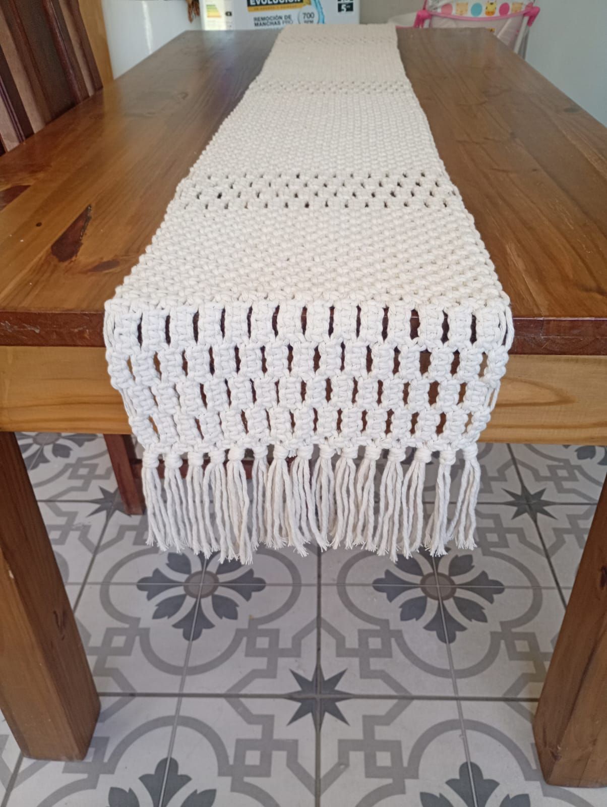 Camino de mesa de macramé color beige sobre una mesa de madera, con flecos en ambos extremos.