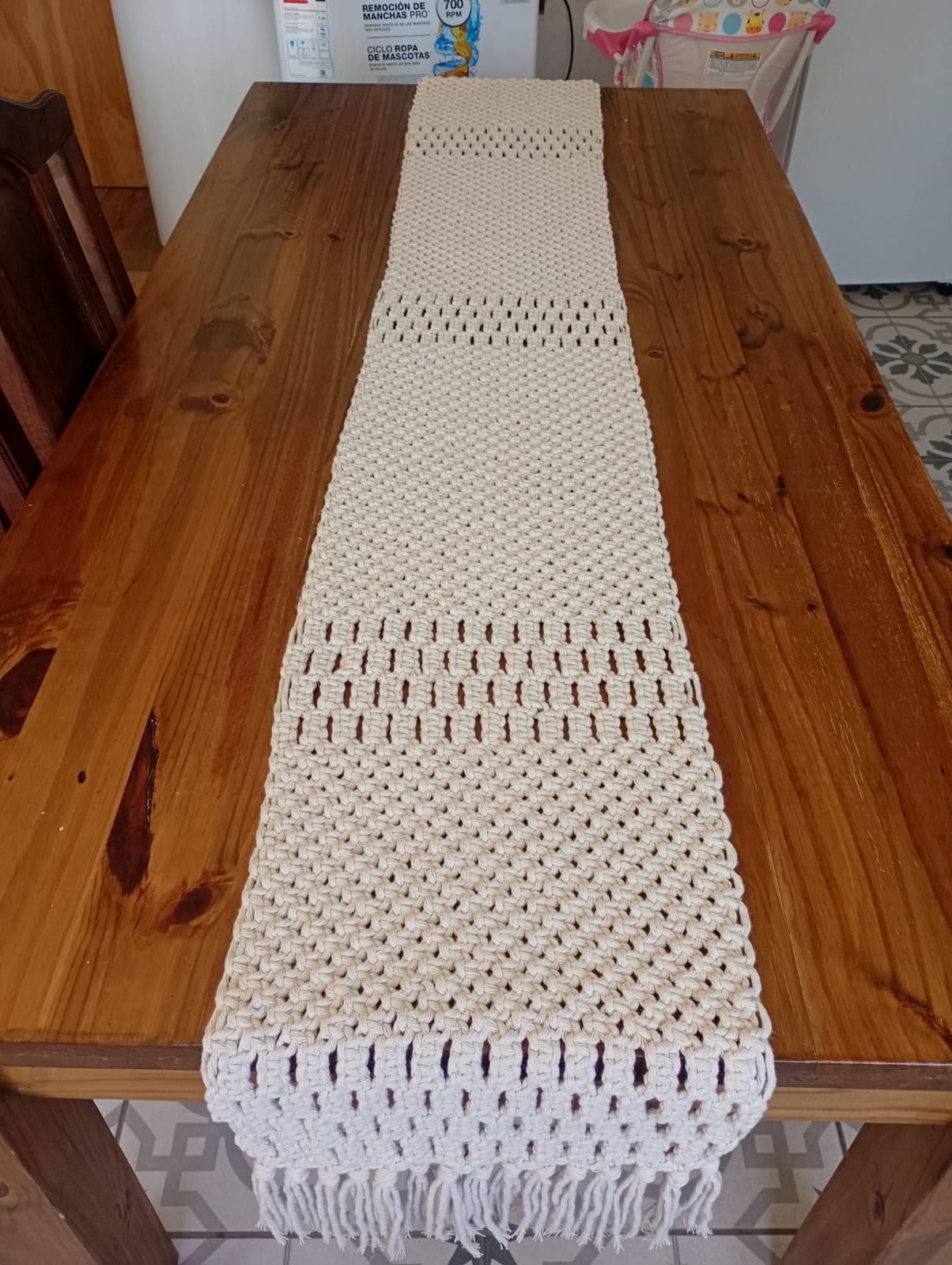 Camino de mesa de macramé color beige sobre una mesa de madera, con flecos en ambos extremos.