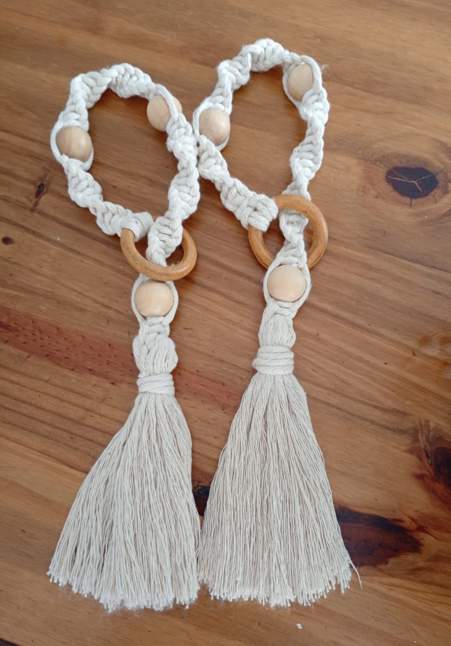 Dos abrazaderas para cortinas de macramé color crema con cuentas de madera y borlas sobre una superficie de madera.