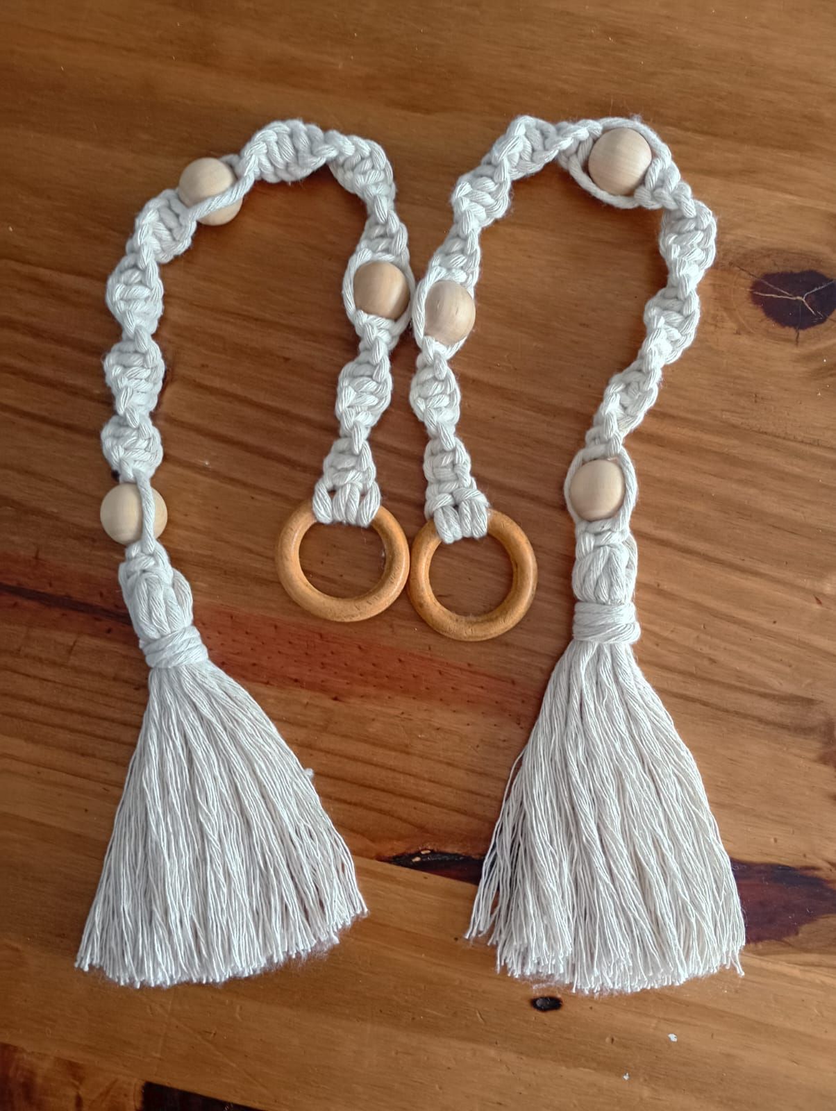 Dos abrazaderas para cortinas de macramé color crema con cuentas de madera y borlas sobre una superficie de madera.