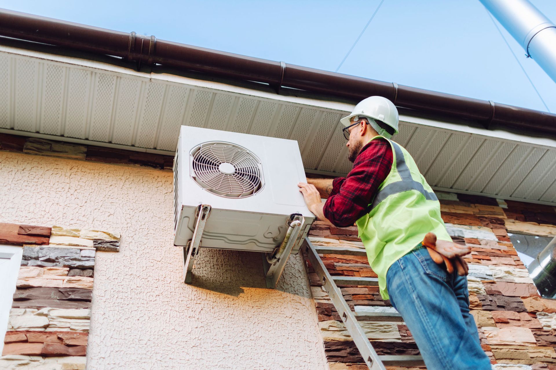 HVAC technician in safety gear installs an air conditioning unit mounted on a house.