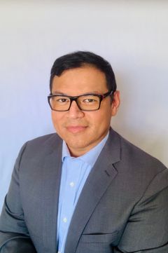 Man in glasses and gray suit, blue shirt, smiles at the camera against a white backdrop.