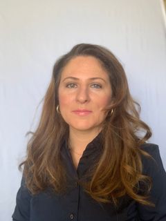 Woman with long brown hair, wearing a black shirt, posing in front of a white wall.