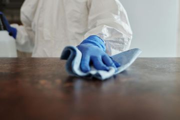 Person in protective suit, blue gloves, wiping a wooden table with a blue cloth.