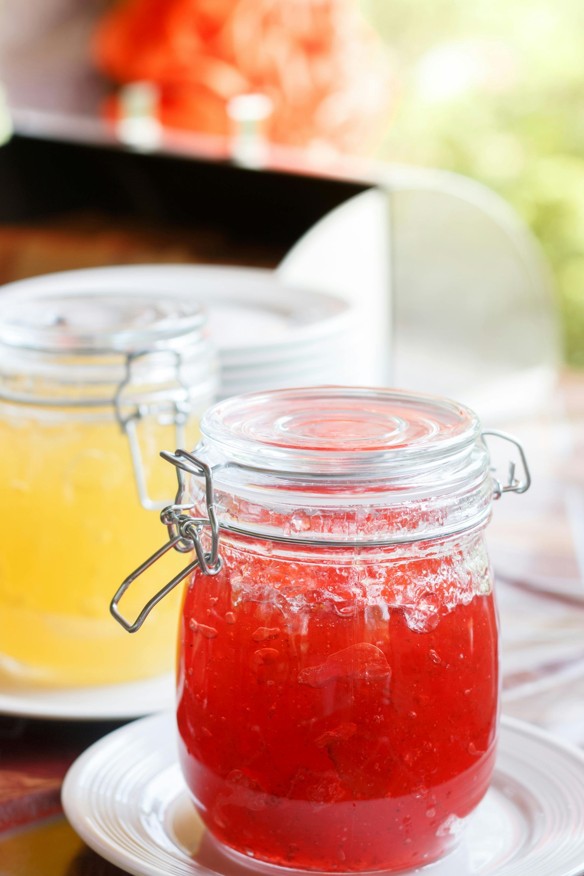 Red and yellow jams in glass jars on white plates, outdoor setting.