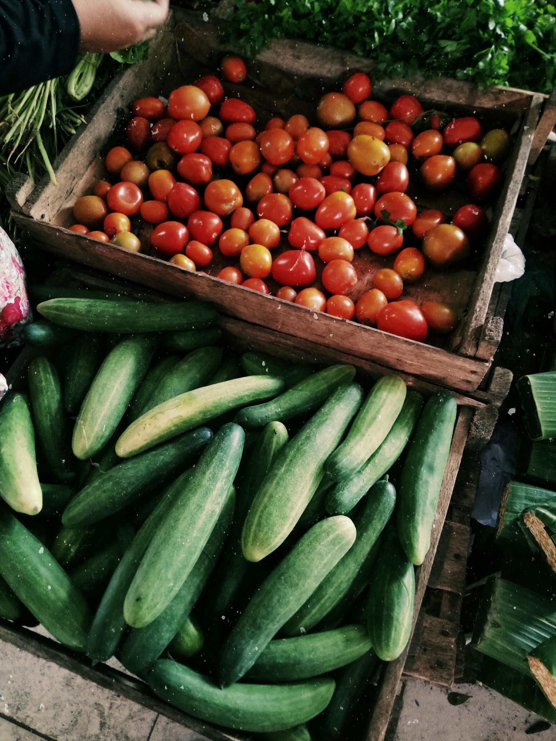 Tomatoes in a wooden crate above a pile of cucumbers at a market stall.