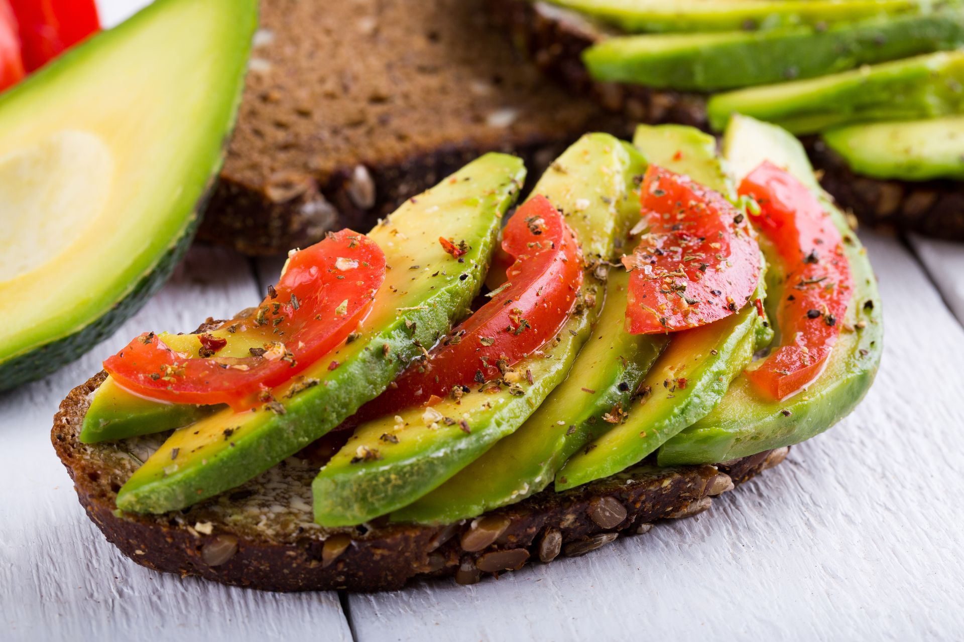 Avocado toast with tomato slices and herbs on dark bread, white wooden surface.
