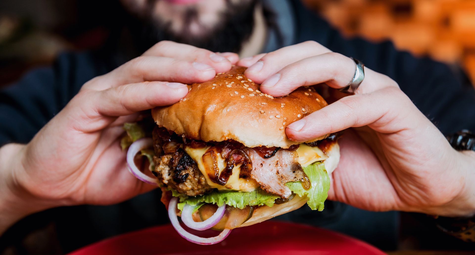 Person holding a large burger with sesame bun, cheese, bacon, lettuce, and onion, on a red plate.