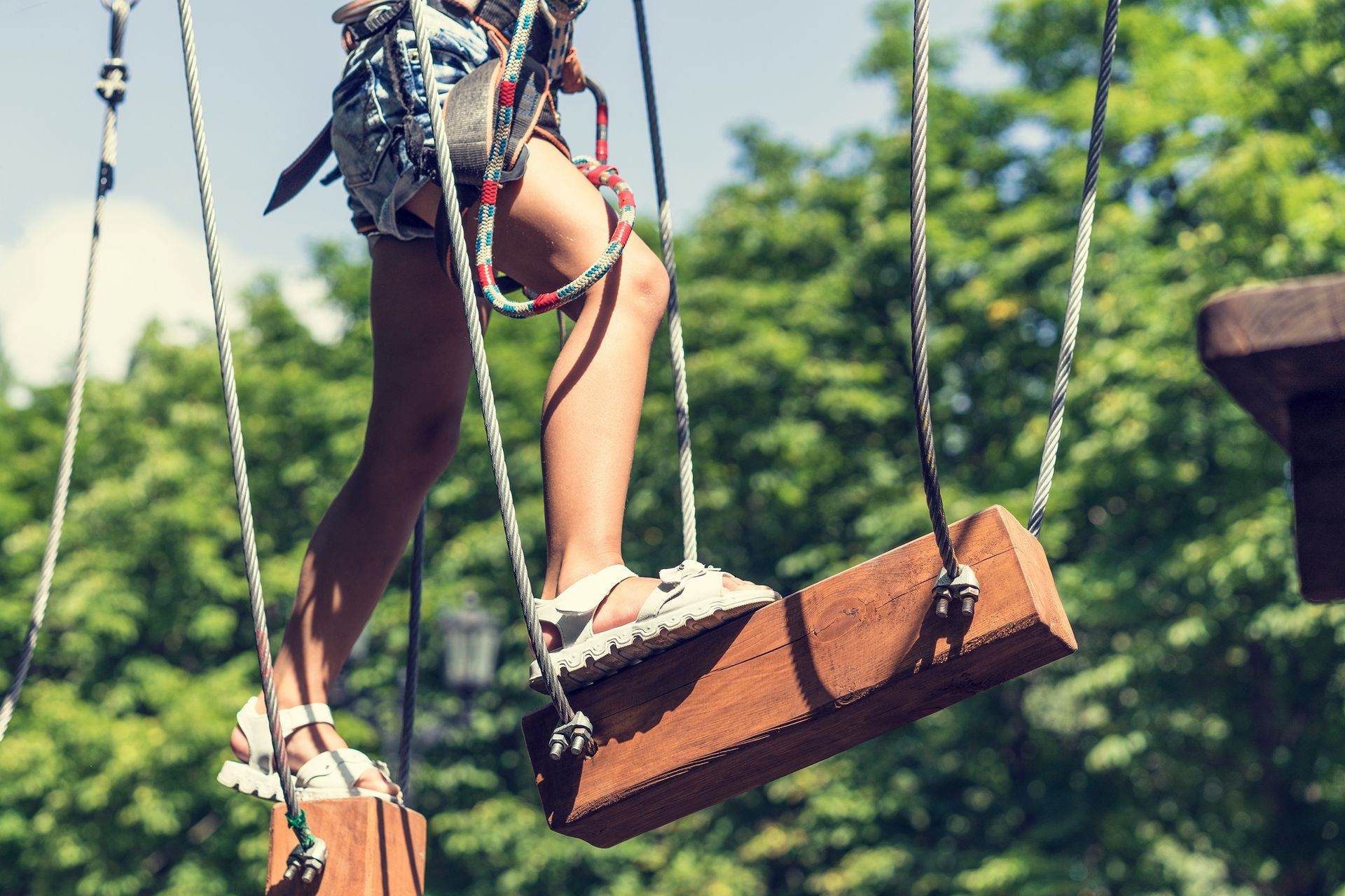 Child in harness walking on a suspended log bridge, outdoors with green trees.