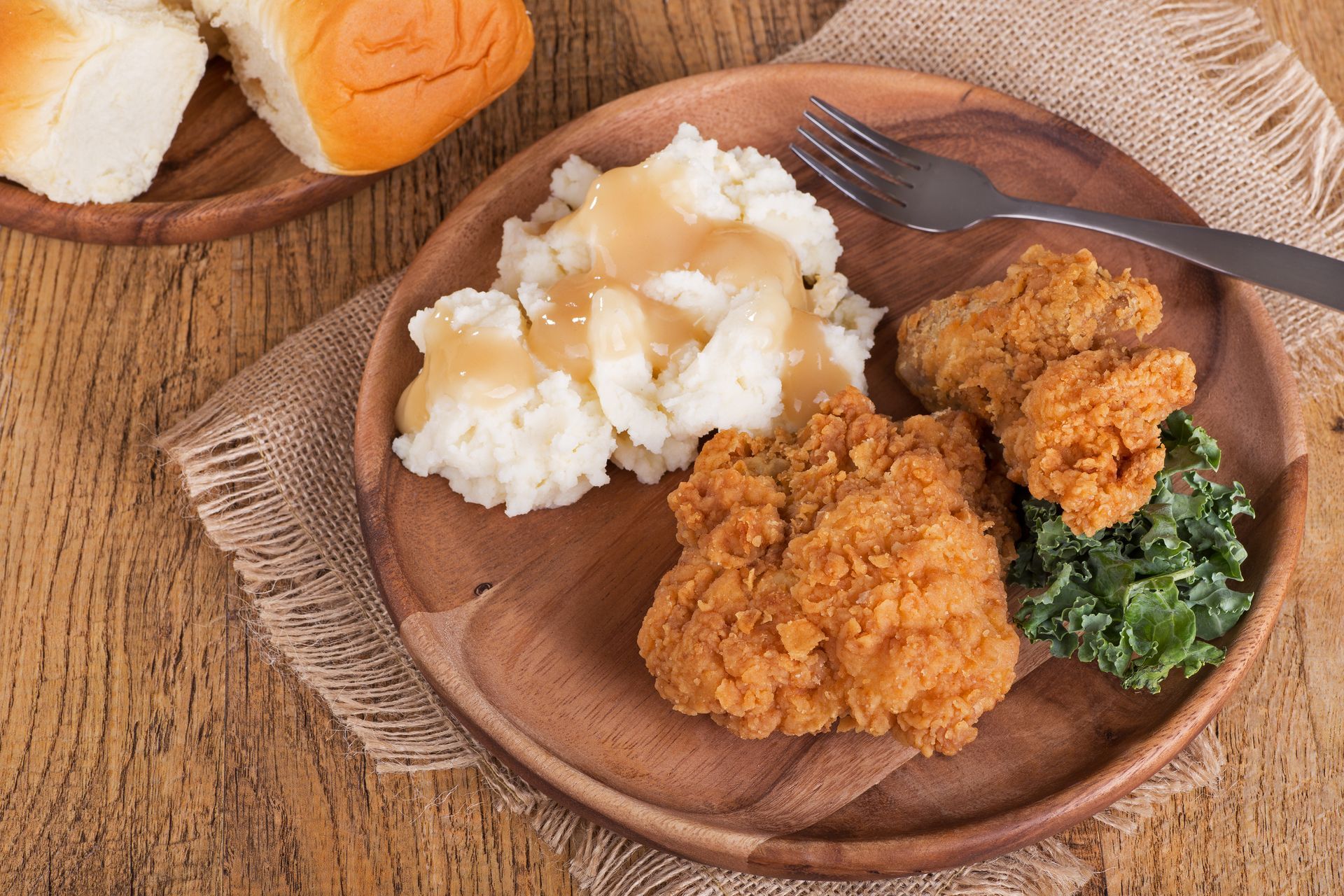 Fried chicken meal: crispy chicken, mashed potatoes with gravy, greens, and rolls on a wooden plate.