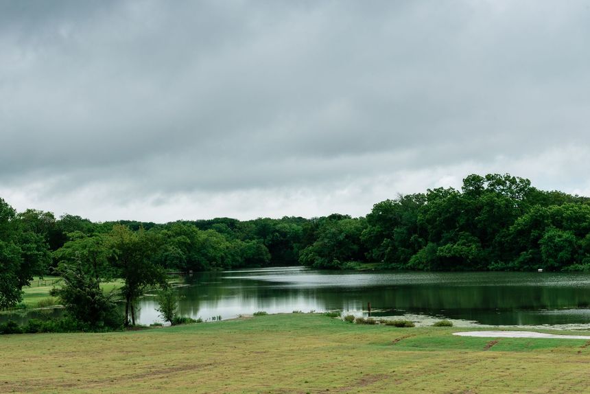 Overcast sky over a calm lake surrounded by lush green trees and a grassy field.