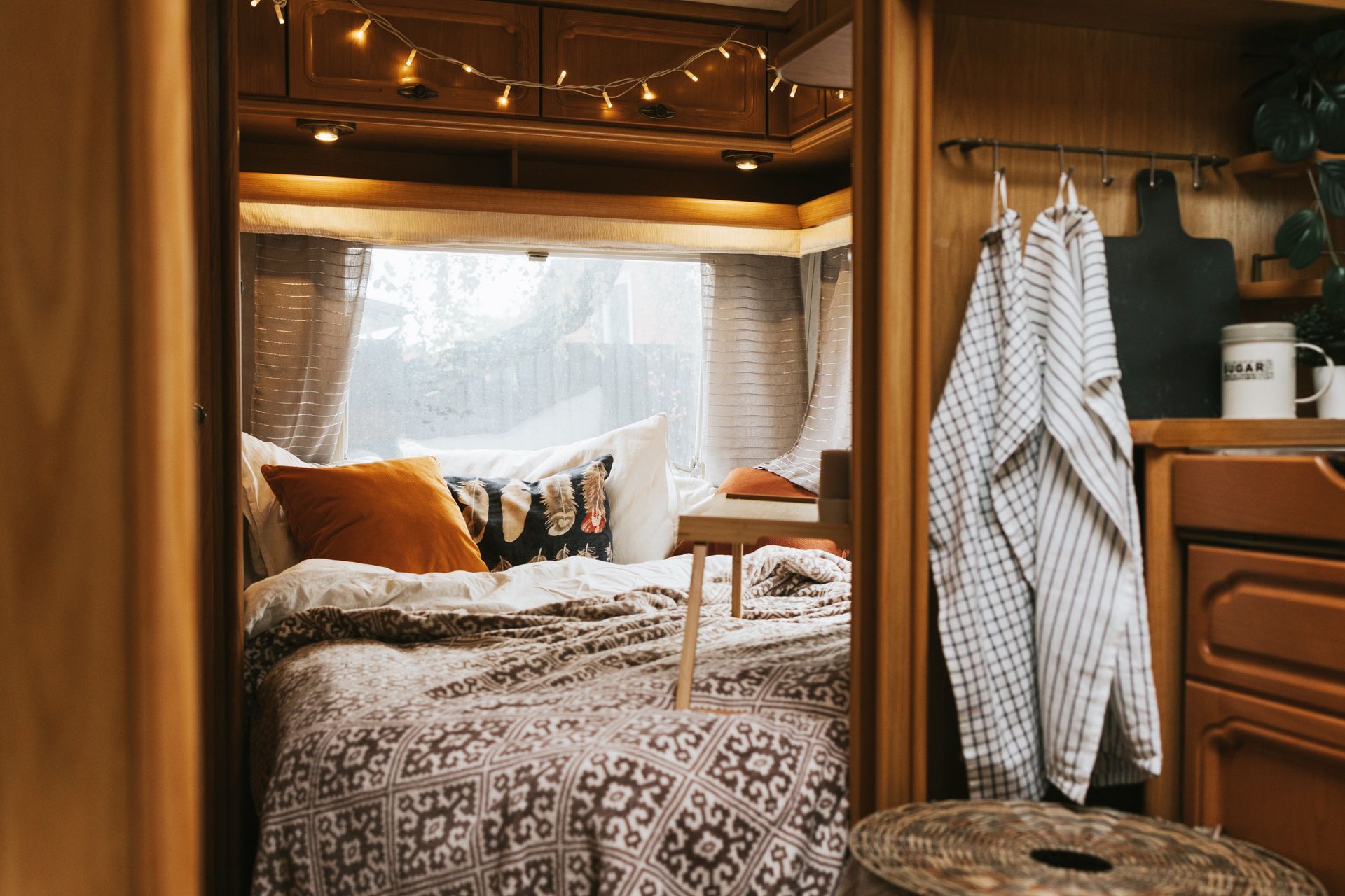 Cozy RV interior with a bed, small table, and decorative lights. Towel and cutting board hang in the kitchen area.
