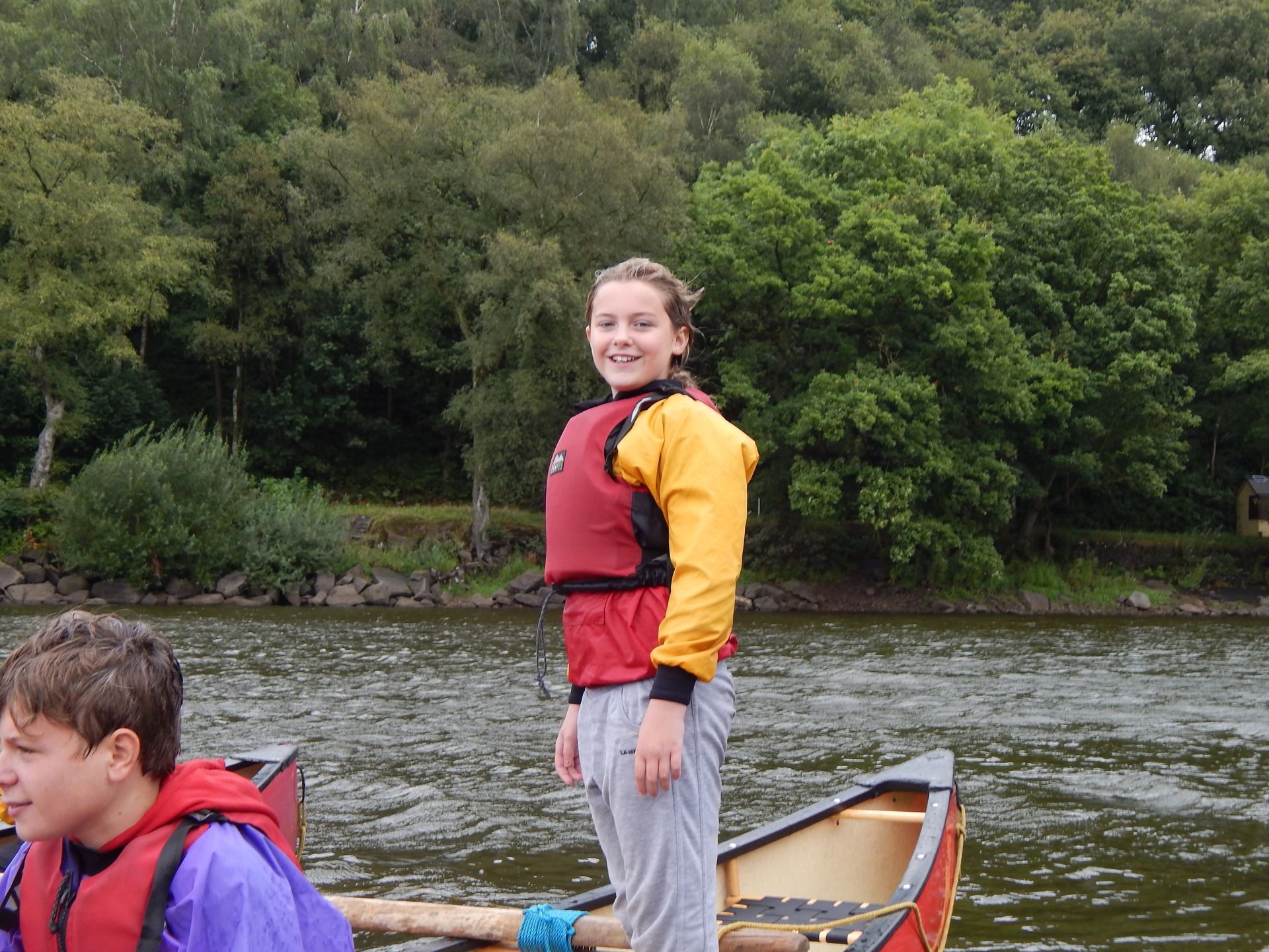 A girl in a life jacket is standing in a canoe on a river.