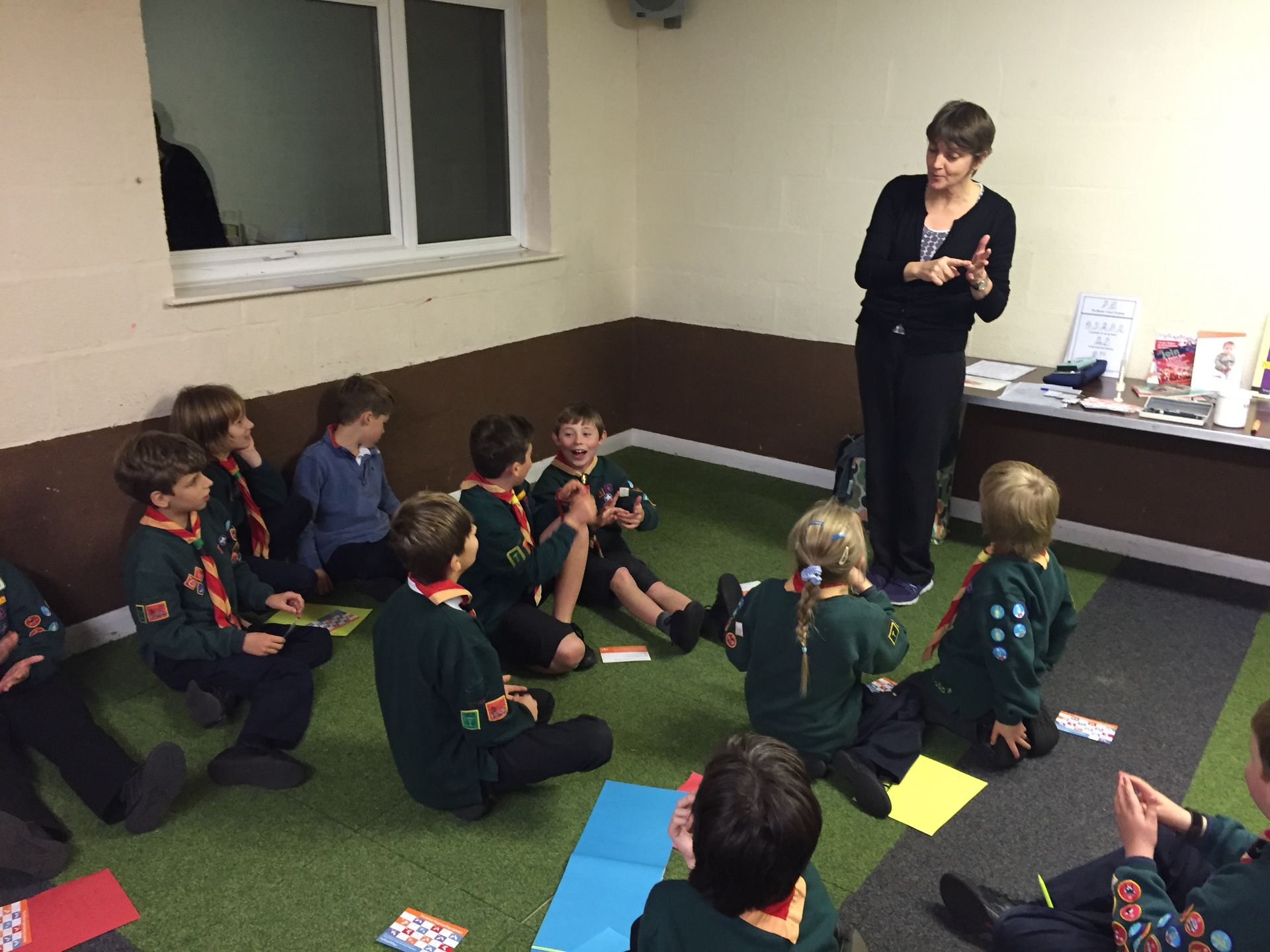 A group of children are sitting on the floor in a room while a woman talks to them.