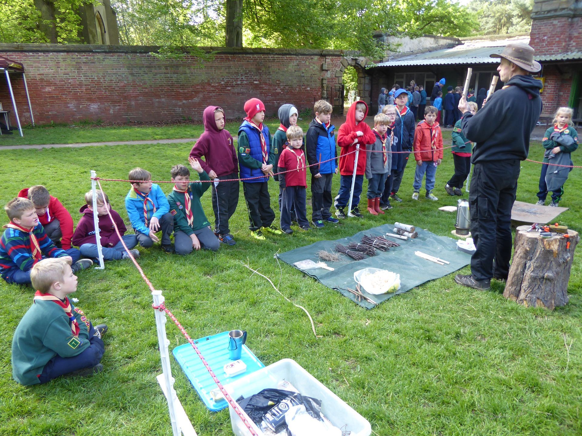 A group of children are sitting on the grass while a man talks to them.