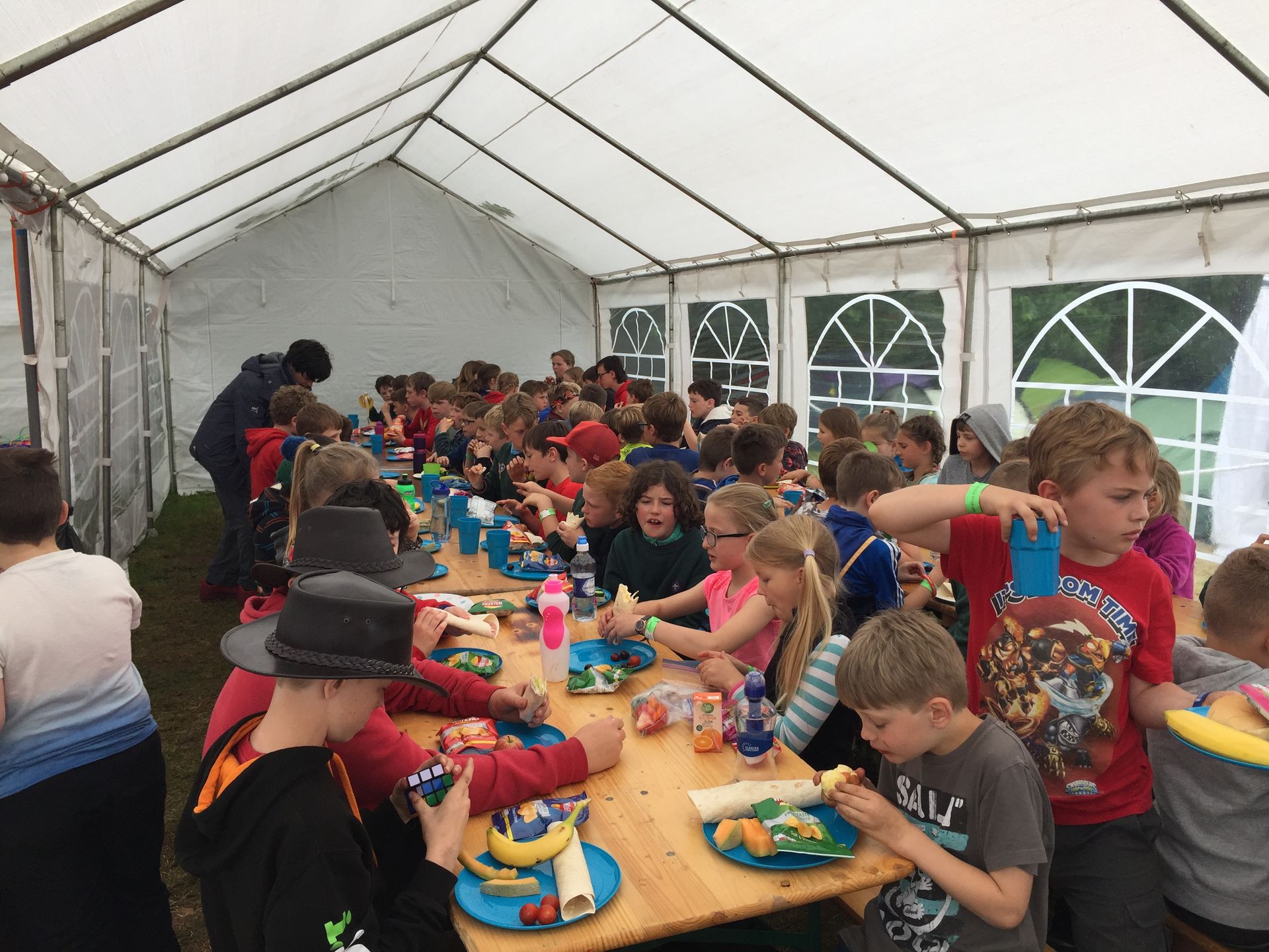 A group of children are sitting at a long table under a tent eating food.