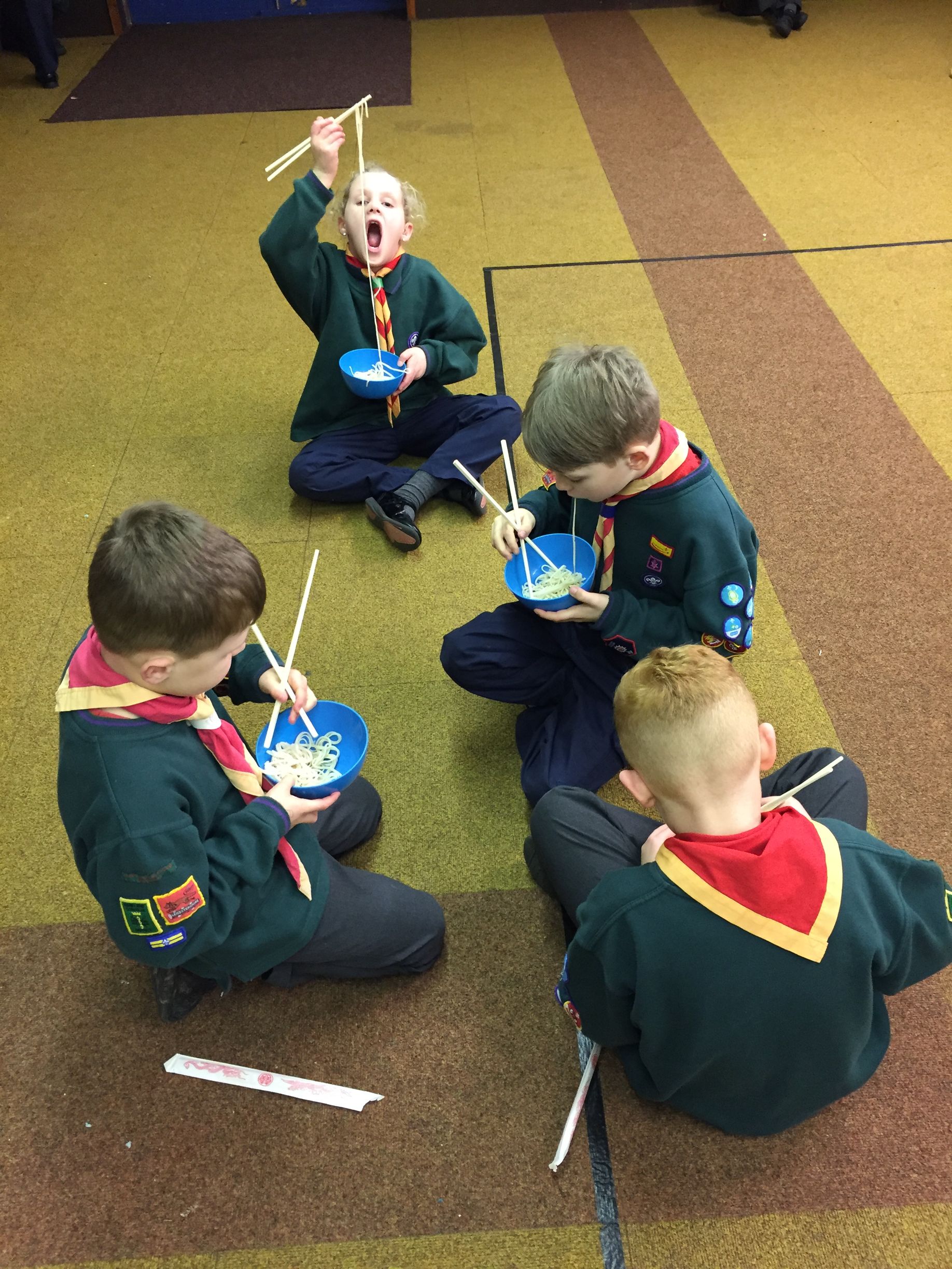 A group of young boys are sitting on the floor eating spaghetti with chopsticks