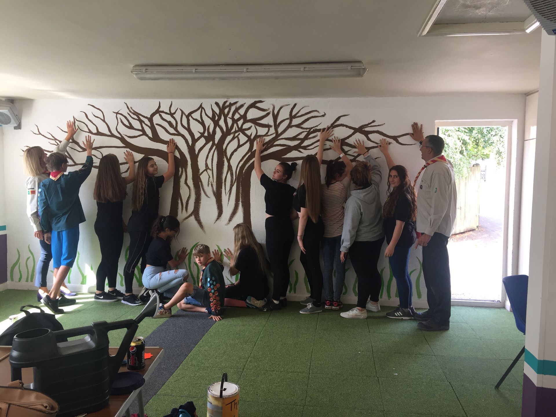 A group of people are standing in front of a wall with a painting of a tree on it.