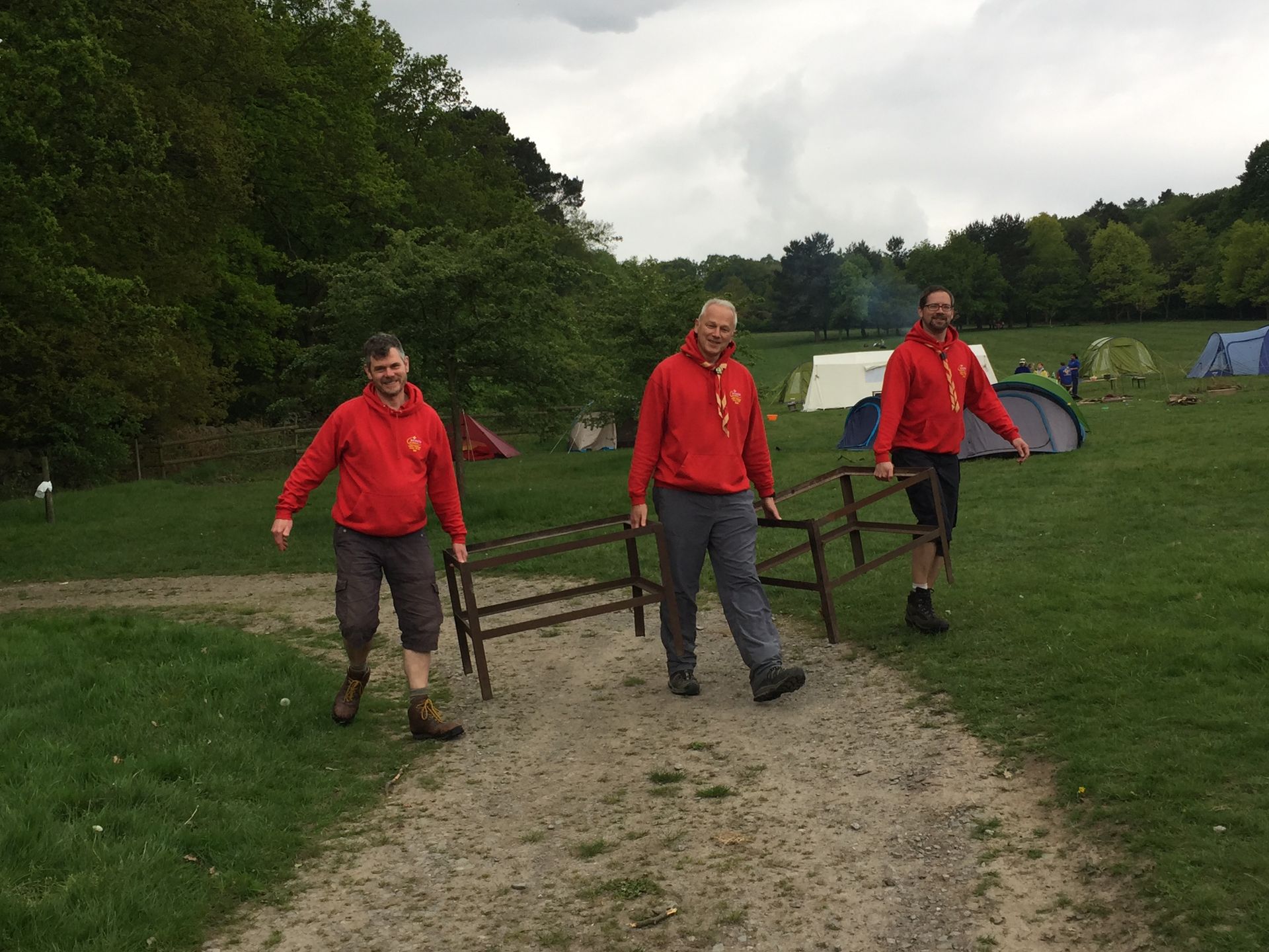 Three men in red jackets are walking down a dirt path carrying a wooden bench.