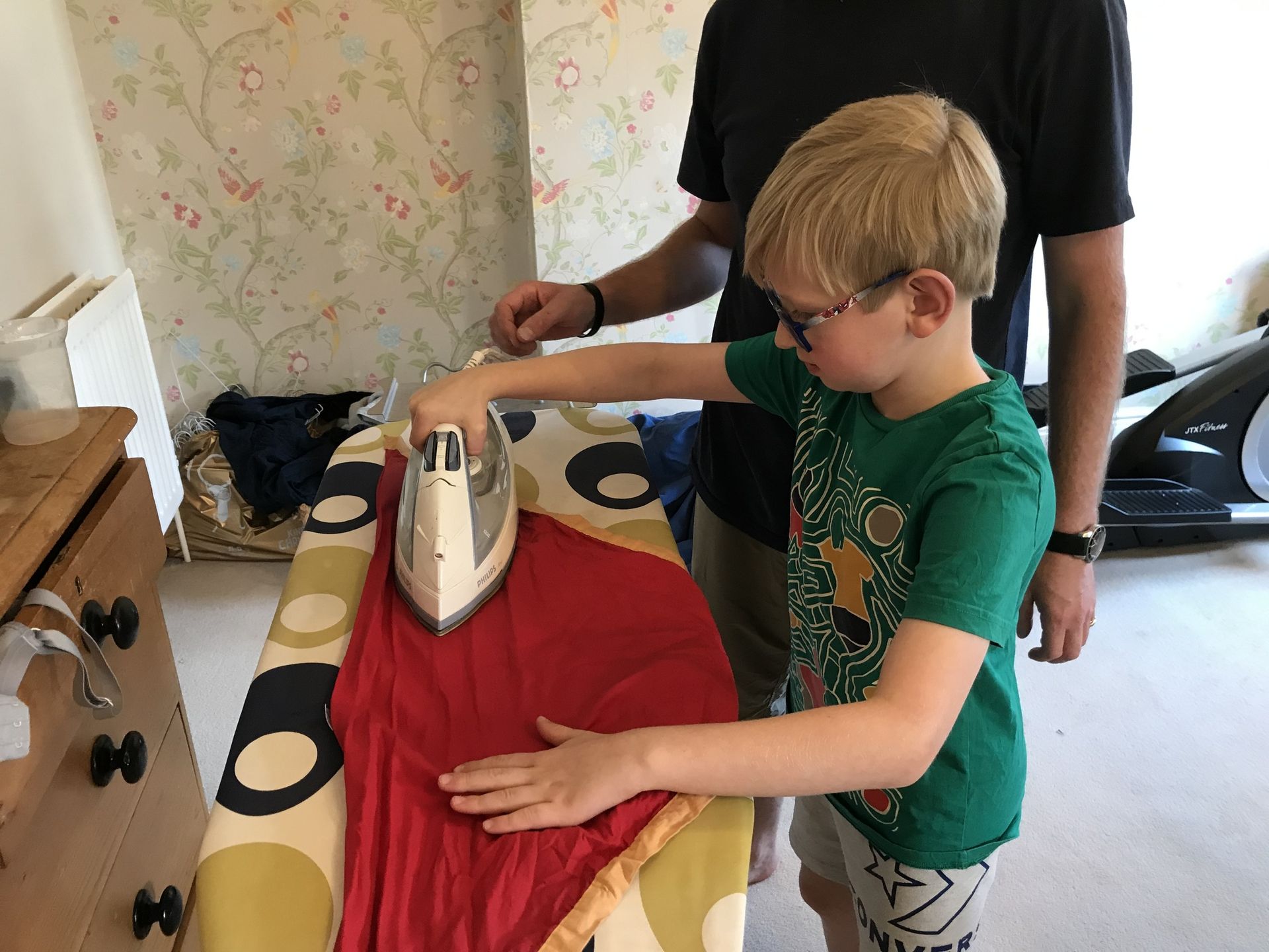 A boy in a green shirt is ironing a red shirt on an ironing board.
