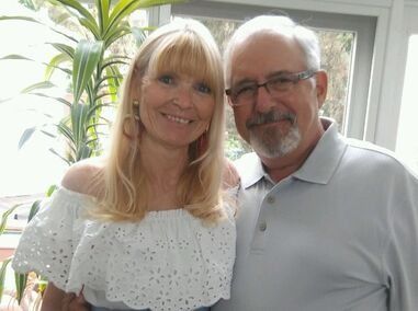 Two smiling adults standing indoors by a window, with a leafy plant in the background