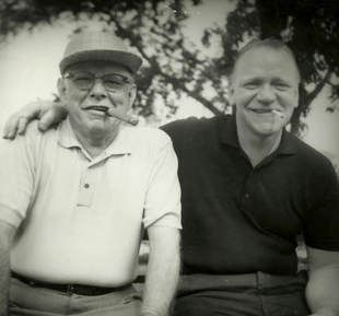 Two men seated outdoors, one with a cigar, smiling with an arm around the other.