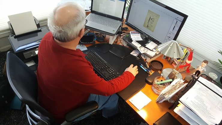 Person in a red shirt working at a cluttered desk with dual monitors and papers.