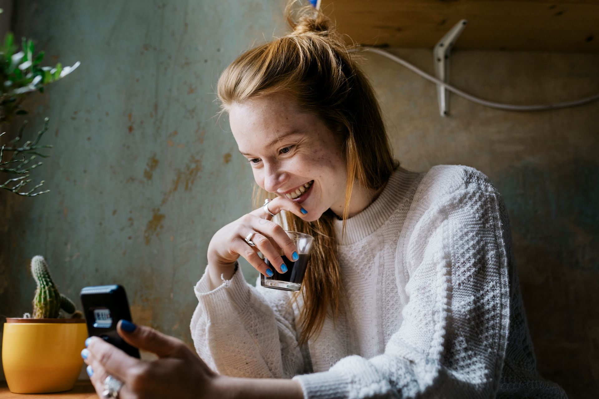A woman is sitting at a table drinking coffee and looking at her phone.