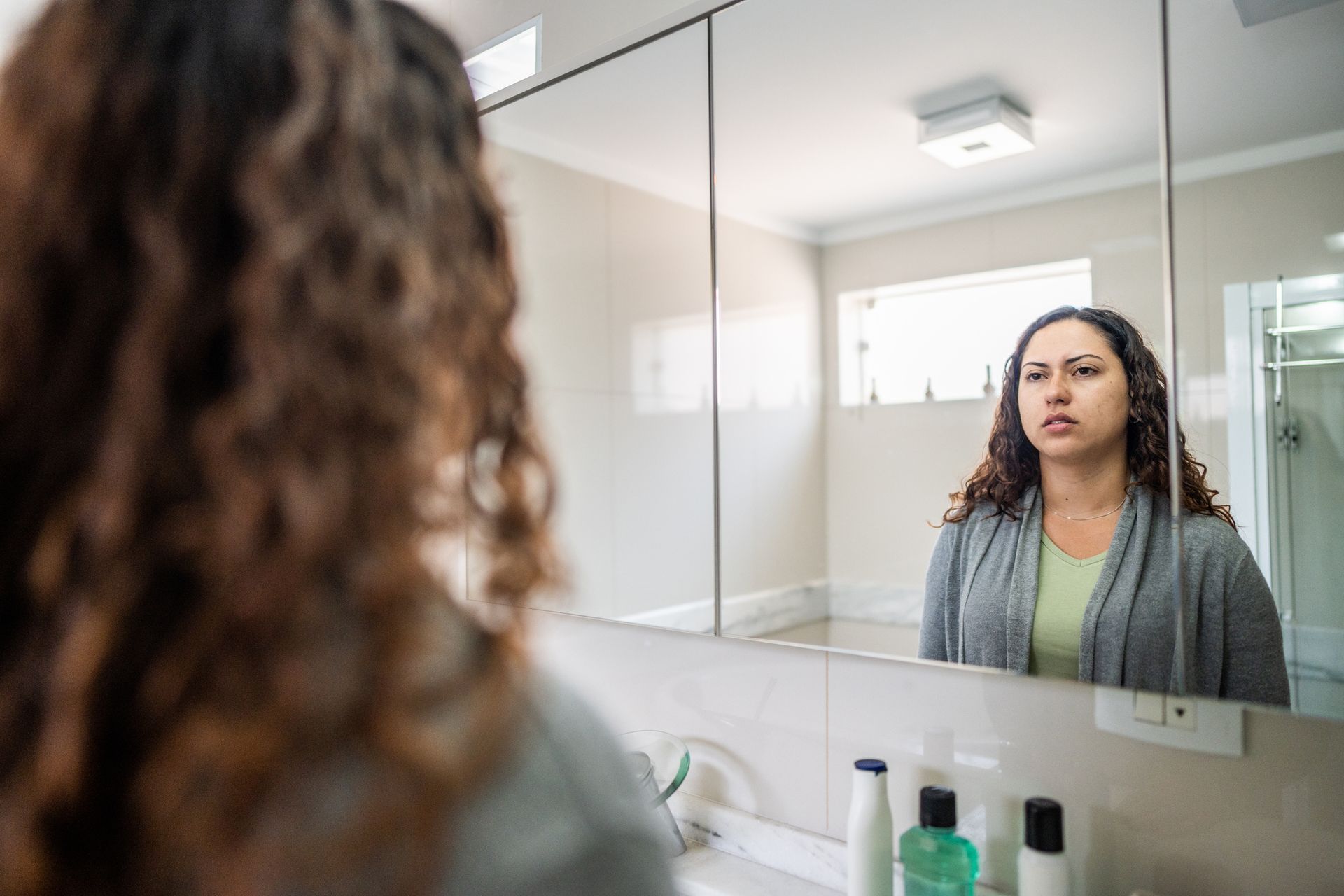 A woman is looking at her reflection in a bathroom mirror.