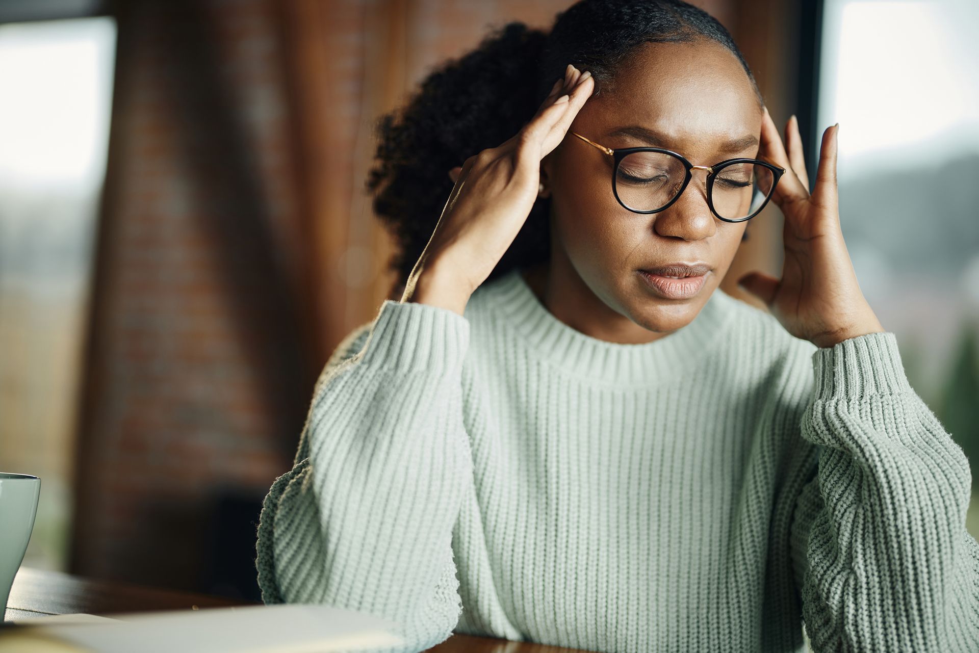 A woman wearing glasses is sitting at a table with her hands on her head.