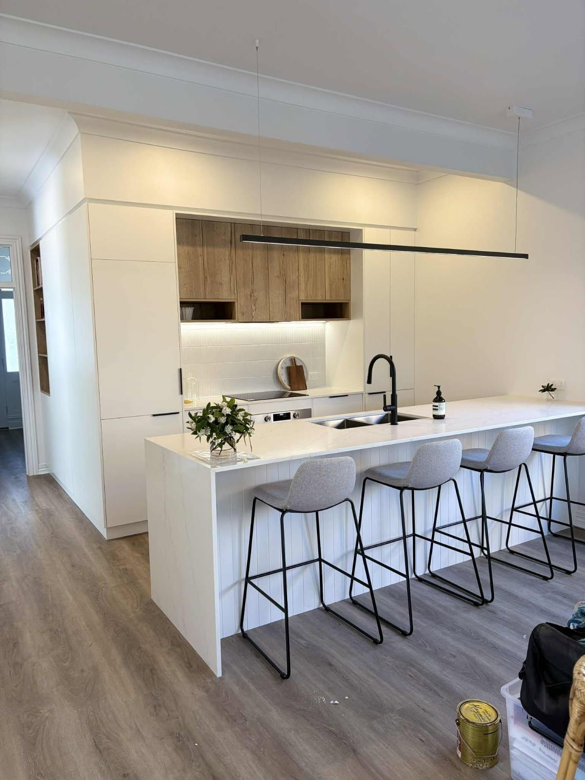 Modern kitchen with white countertops and cabinets, wood accents, black faucet, and gray bar stools.