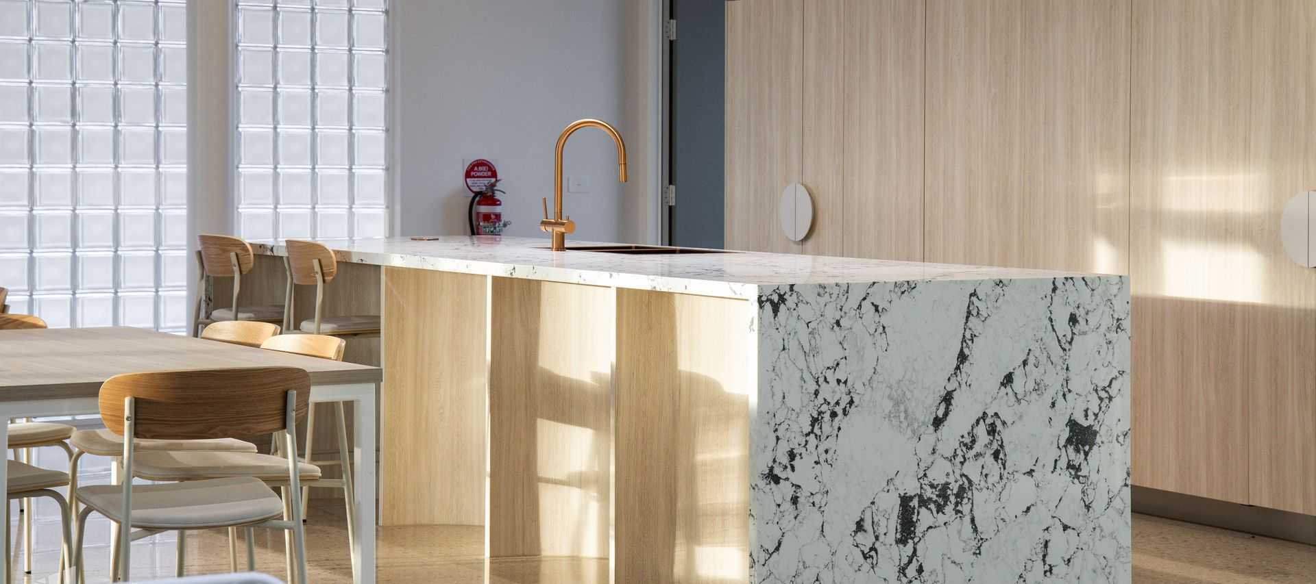 Kitchen with island. Marble countertop, copper faucet, light wood cabinetry. Glass block window behind.