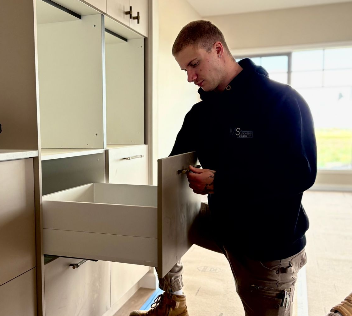 Man installing a drawer in a built-in cabinet. He wears a black hoodie and tan work boots in a room with a window.