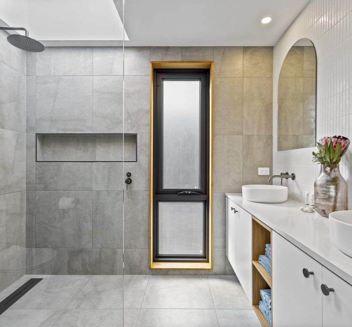 Modern gray tiled bathroom with shower, long window, vanity, and large arched mirror.