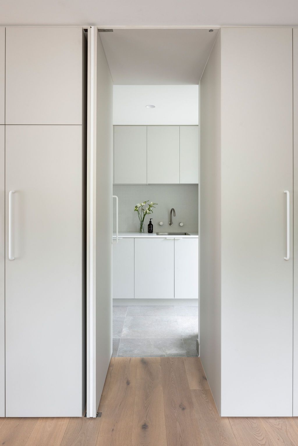 Sliding white door open to reveal a light-filled kitchen with white cabinets, countertops, and a sink.