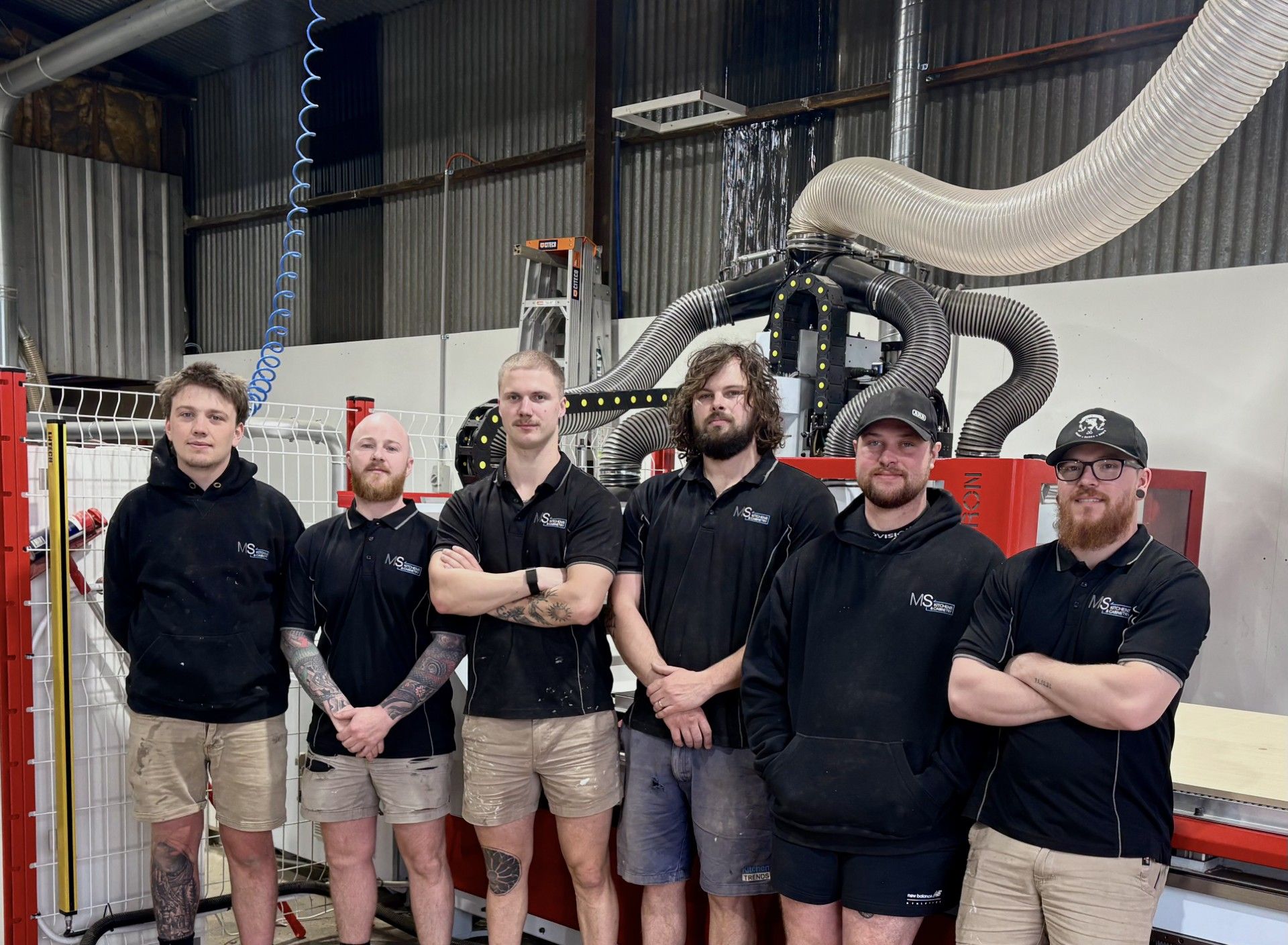 Six people standing in front of industrial machinery. They wear work clothing and look at the camera. A workshop setting.